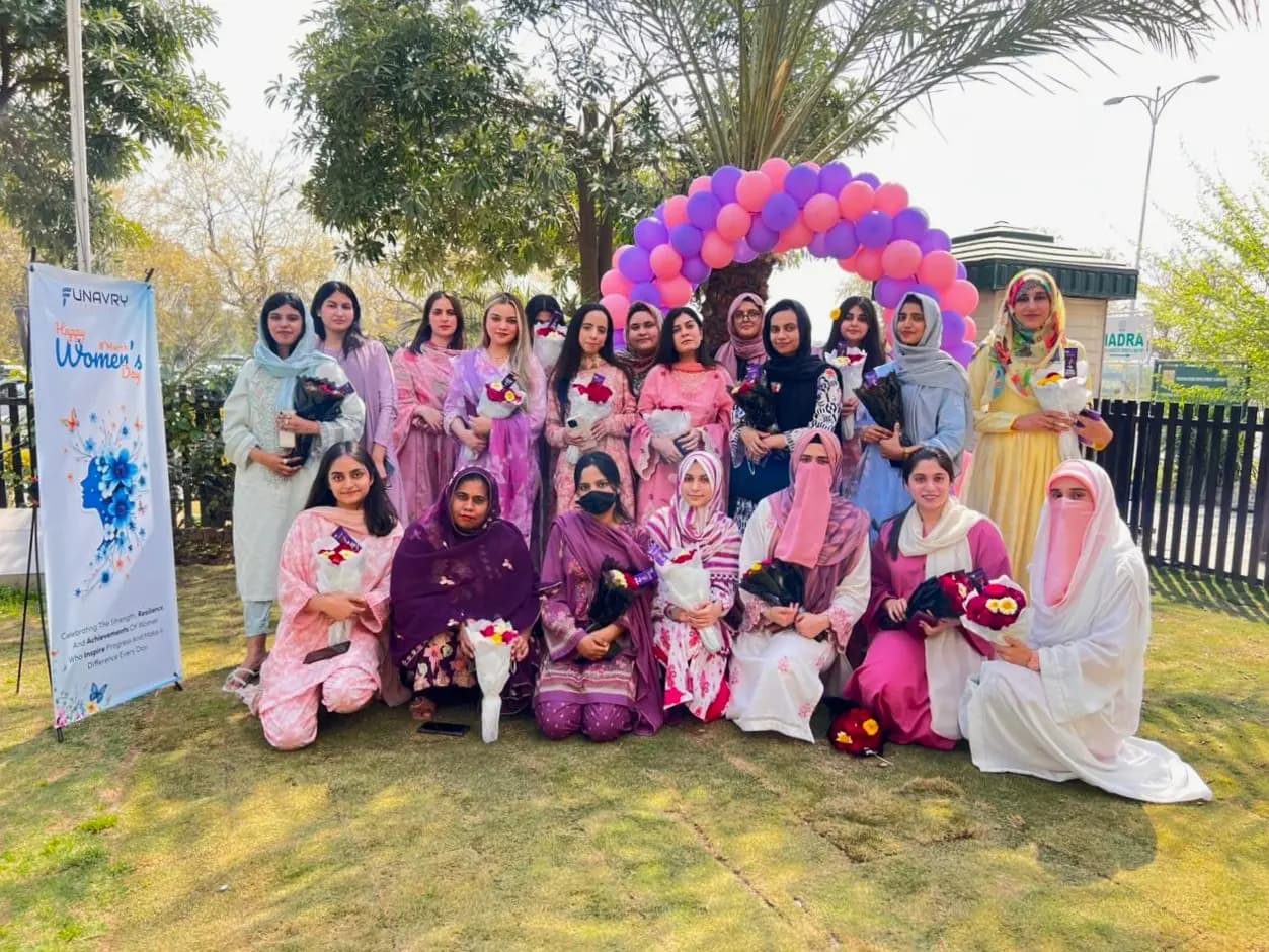 Women of the team holding bouquets under a balloon arch beside a Happy Women's Day banner